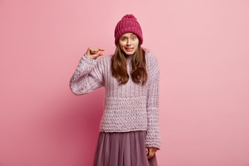 I need little bit. Puzzled lady shows very tiny object, shapes something small, wears headgear, knitted jumper and skirt, looks in puzzlement, isolated over pink background, gestures indoor.