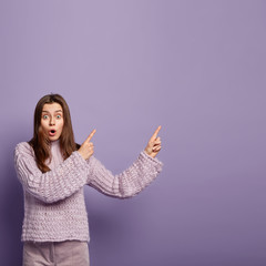 Photo of surprised European woman has shocked expression points aside with both fore fingers, opens mouth from wonder, demonstrates something amazing, wears knitted jumper, isolated on purple