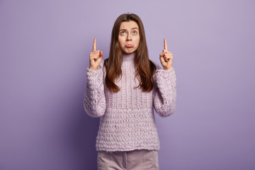 Photo of upset gloomy woman with miserable facial expression, points upwards, purses lips, feels...