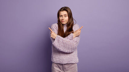 Photo of hesitant woman with puzzled facial expression points sideways, indicates two objects on right and left, cant make choice, faces doubtful situation, has unsure look, wears oversized jumper