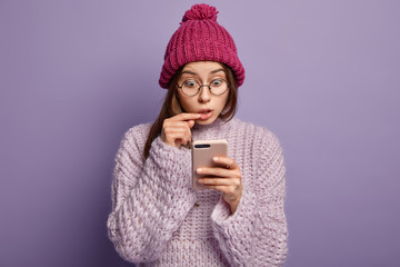 Headshot of astonished lady checks mailbox, focused in screen of cell phone, has conversation in group chat, looks surprisingly at sms, wears winter hat and sweater, isolated over purple background
