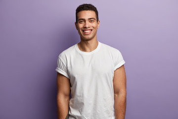 Waist up shot of handsome cheerful man with toothy smile, muscular arms, being in high spirit as spends free time with friends or girlfriend, wears casual white t shirt, isolated over purple wall - obrazy, fototapety, plakaty