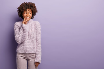 Photo of joyful woman has dreamy cheerful expression, looks away, keeps fore finger near lips, dressed in knitted jumper and trousers, stands in studio against purple background with free space aside