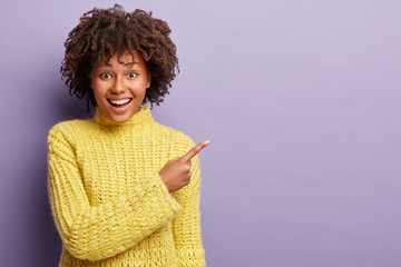 Happy positive woman with Afro haircut, pleased expression, shows direction to beauty salon, enjoys advertising product, wears yellow clothes, isolated over purple background. Promotion, ethnicity
