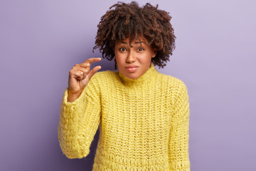 Size matters. Puzzed pretty young woman with Afro hairstyle, has tiny object, demonstrates measure symbol, shapes something small, wears knitted yellow sweater, isolated over purple background
