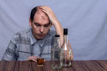 A man sits at the table and looks at a glass with alcohol. Before him stands two empty bottles of alcohol.
