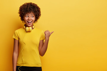 Waist up shot of African American hipster girl being amused by listening music, has modern headphones on neck, points aside, shows way to electronic shop, has cheerful expression, isolated on yellow