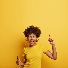 People, life, energy and music concept. Glad woman rejoices something, enjoys nice sound in new headphones, uses smartphone, listens song from playlist, spends time for dancing and entertaining