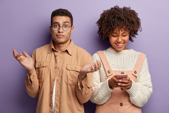 Indoor shot of pleased Afro American woman reads text message on cellphone, dressed in white knitted jumper and pink overalls, hesitant young guy spreads hands, doesnt know what to talk about