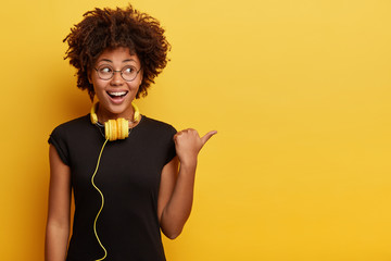 African American woman in high spirit points away with thumb, feels entertained and satisfied, listens radio broadcasting with modern headphones connected to telephone, checks sound, shows free space