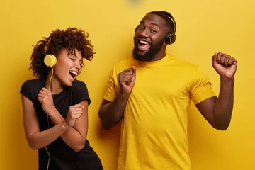 Fototapeten Cheerful young African American bloggers enjoy favourite playlist in headphones, listens audio in music app, feels happiness, dance actively against yellow background, move emotionally, have fun © Wayhome Studio