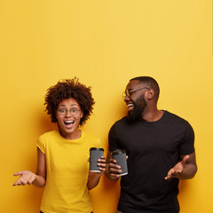Positive black classmates have coffee break, hold paper cups, discuss future plans with happy expressions, share good news, stand shoulder to shoulder over yellow background, free space above