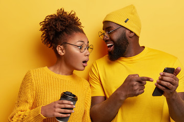 Surprised curious dark skinned woman looks at screen of boyfriends cellular, reads online post with...