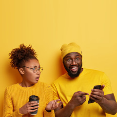 Satisfied cheerful black man points at screen of cell phone, shows funny photos to girlfriend, demonstrates meme, connected to wireless internet, puzzled woman gestures actively and holds coffee