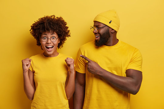 Satisfied Black Bearded Man Points At Cheering Successful Afro American Woman Who Raises Clenched Fists, Achieved Something Desirable, Wear Casual T Shirt, Stand Closely Against Yellow Background.