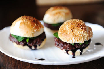 Three freshly cooked burgers on a white plate. Sesame bun, beef cutlet, spinach greens and flowing berry sauce.