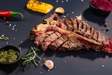 Set of grilled sliced T-bone steak with grilled corn, sauce, chilli, rosemary, salt, garlic on black background.