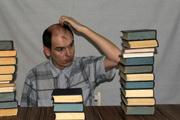 A man sits at a table on which there are piles of books. A man looks at one pile of books and...