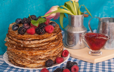 Pancakes on a platter with raspberries, blueberries and blackberries on a kitchen towel. A jug of spring flowers tulips. Berry jam in a transparent vase. Lots of pancakes stacked. Thin pancakes with c