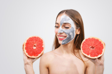 Cheerful young female model has cosmetic cleansing mask on face looking on the grapefruit