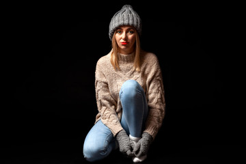 Young beautiful blonde girl in knitted gloves and hat in gray, blue jeans, beige sweater sits on the floor with crossed legs and looking to the camera on a black isolated background