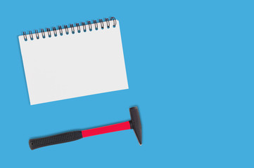 Blank paper notebook with spiral holder near hard metal hammer lies on blue table in workroom. Top view. Repair concept