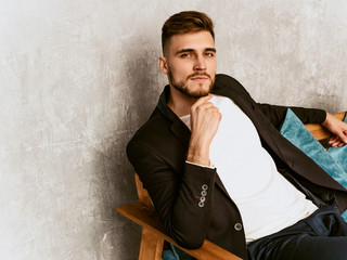 Portrait of handsome hipster lumbersexual businessman model wearing casual black suit. Fashion stylish man posing sitting in chair in luxury interior. Thinking