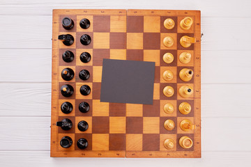 Classic wooden chess board with chess pieces. Chess set on game board with blank paper card, top view. Competition and business concept.
