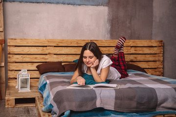 Beautiful girl in red pants and socks is lying on the bed reading a book, magazine. Hobby, free time, leisure. Space for text. The gaze is directed down into the book.