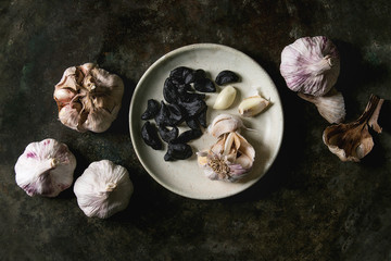 Variety of fresh organic garlic bulbs whole and peeled and cloves of black fermented garlic on ceramic plate over dark metal background. Flat lay, space