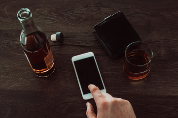 A man presses a finger on a mobile phone. Next on the table is a glass of whiskey, a bottle of whiskey and a purse.