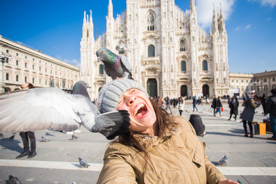 Winter travel, vacations and birds concept - Young happy woman tourist with funny pigeons making selfie photo in front of the famous Duomo cathedral in Milan.
