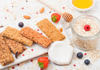 Organic cereal granola bar with berries on marble board with honey spoon and jar of oats and coconut on marble background.