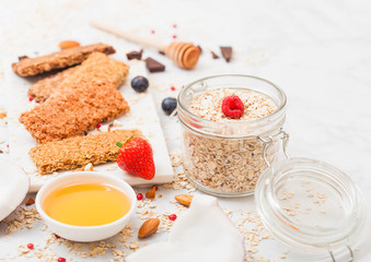 Organic cereal granola bar with berries on marble board with honey spoon and jar of oats and coconut on marble background.