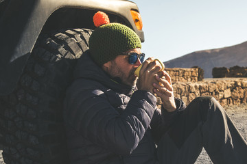 Bearded middle-aged man with woolen hood and sunglasses resting sitting after a trekking day Male...