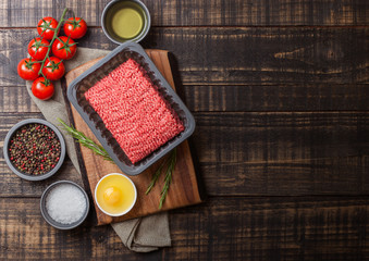 Tray with raw minced homemade beef meat with spices and herbs. Top view and space for text. On top of wooden kitchen table background. With pepper salt and tomatoes.