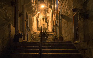 Night view on a alleyway goes upstairs, lights and handrail covered by old house fronts. Some flowers in the pot. Clothes hanging down in the background. Located in Malta.