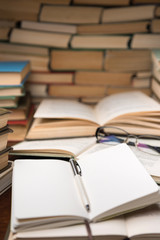 Education learning concept with open book and notebook on the table. Stack piles of books on reading desk and glasses in bookshelves background