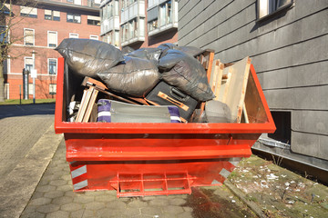 Full orange metal Skip container standing on the road near the house. In the container are old...