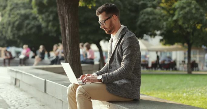 Young attractive caucasian businessman sitting at the bench at the city park and working on his laptop computer.