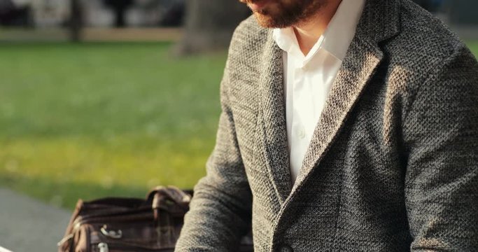 Close up of the rich Caucasian young businessman typing on the laptop computer while sitting outdoors in the park on the bench.