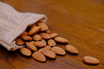 Almonds in a small sack poured out on wooden table shot under warm lighting.