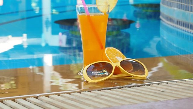 Glass of orange juice with orange slice and orange sunglasses at outdoor swimming pool. Tilt up shot.