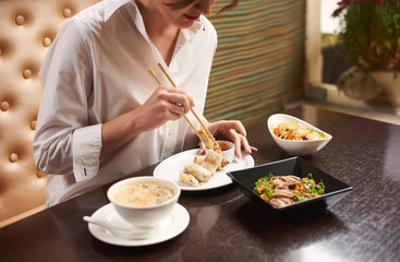 Brunette woman in white blouse with red lips ordering meal in luxury Japanese restaurant. Girl eating four course lunch including soup, tempura shrimps, noodles and warm salad using food sticks.