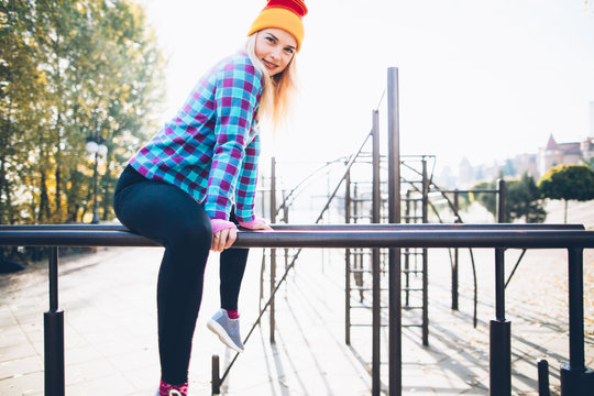 Young beautiful woman relaxing during street workout sitting on parallel bars at calisthenics park, looking at camera