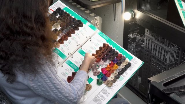 The girl in the hairdresser chooses the color of paint for hair coloring. She sits in front of the mirror and leafs through a catalog of paint samples. Rear view from above. 4k. 25 fps.