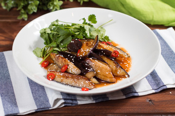 Eggplant slices in spicy red sauce in clay bowl on wooden background. The view from the top.
