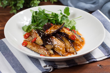 Eggplant slices in spicy red sauce in clay bowl on wooden background. The view from the top.