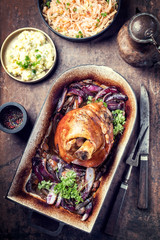 Traditionelle gegrillte bayrische Schweinshaxe mit Zwiebeln, Kartoffelsalat und Sauerkraut als Draufsicht in einem rustikalen Schmortopf auf einem alten Board