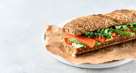 sandwich from a cereal baguette with avocado, salmon, cream cheese, tomatoes and lettuce leaves on a white plate.  light background, selective focus and copy space